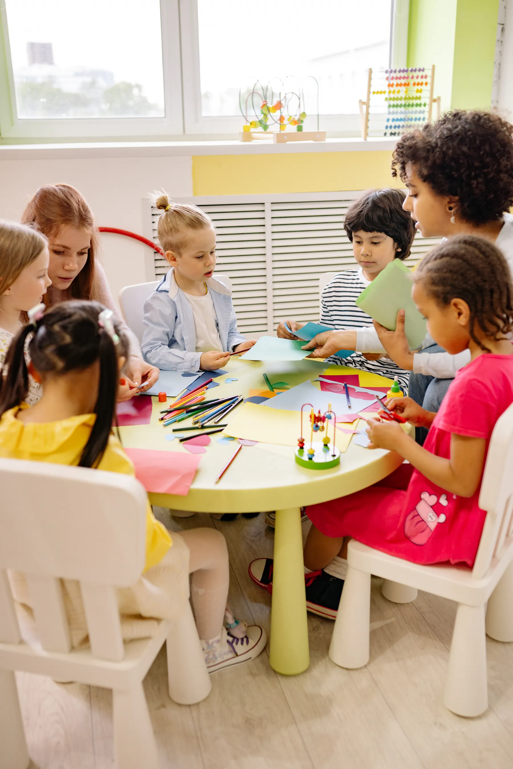 Children making art at a yellow classroom table with an adult alongside